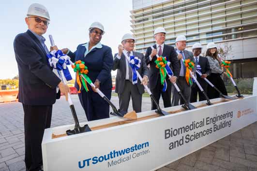 People wearing hard hats dig with shovels inside a sandbox printed with the UT Southwestern and UT Dallas logos and the words Biomedical Engineering and Sciences Building