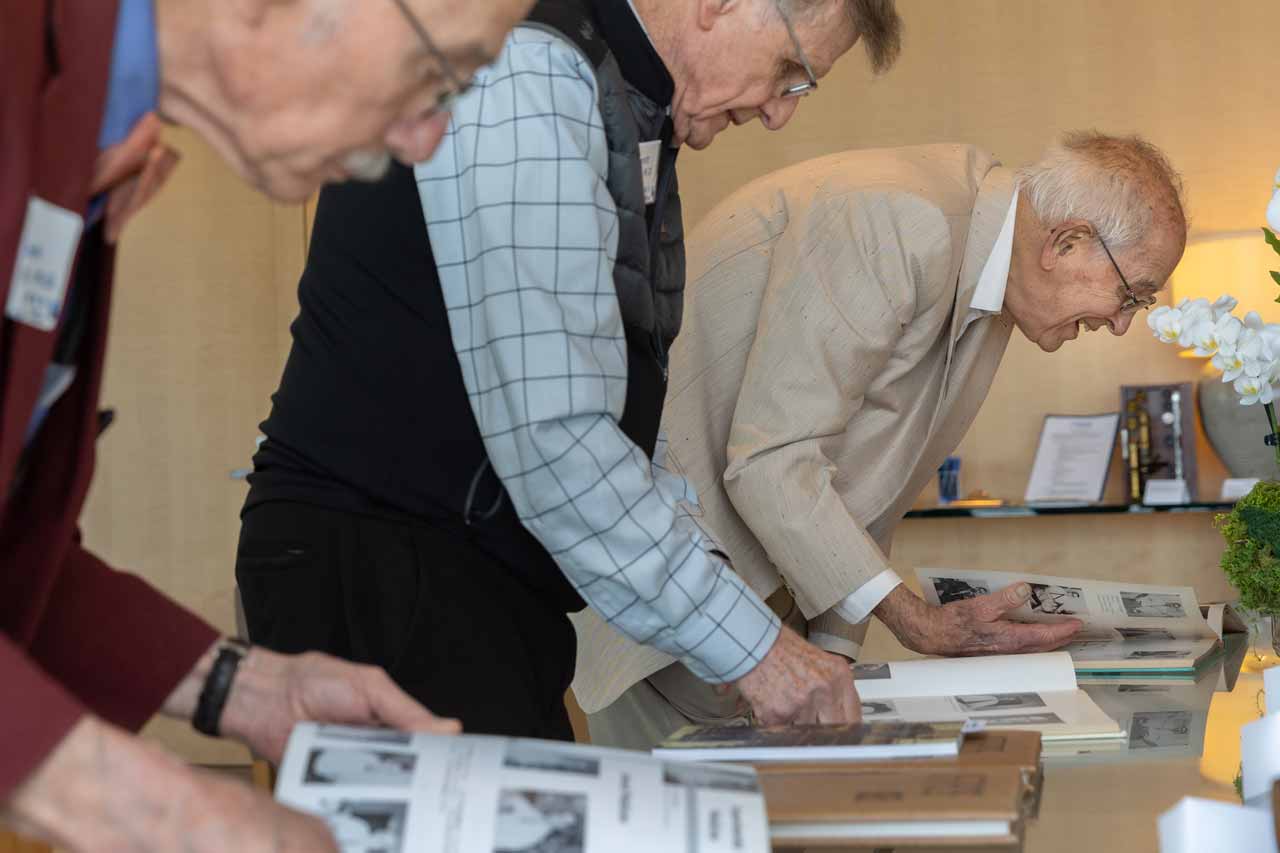 A row of three older men lean over a table to look closely at three yearbooks.