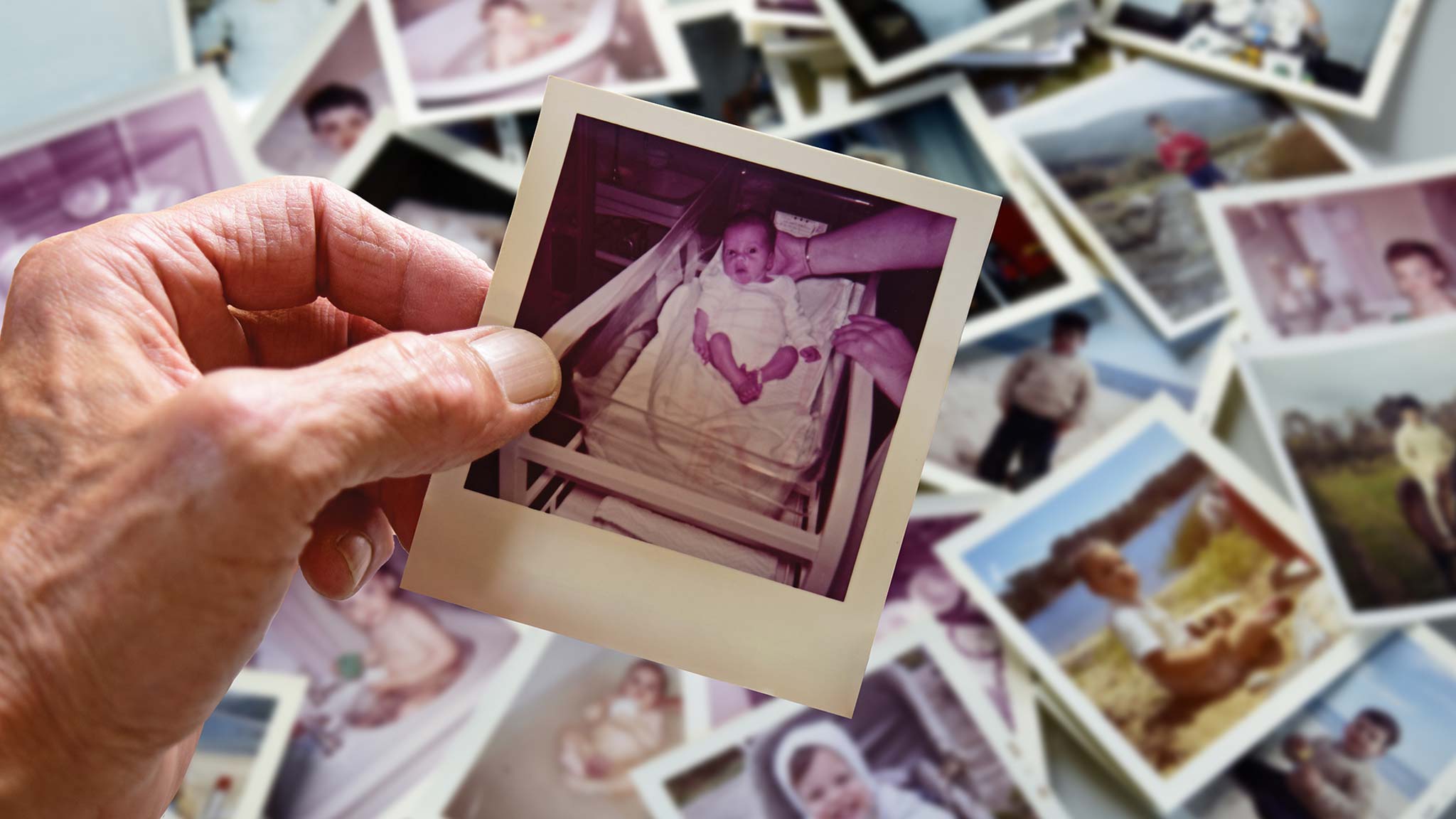 An image of an elderly hand holding a photograph of a baby in a hospital bassinet. Behind the hand, a collection of family photos is strewn across a countertop.
