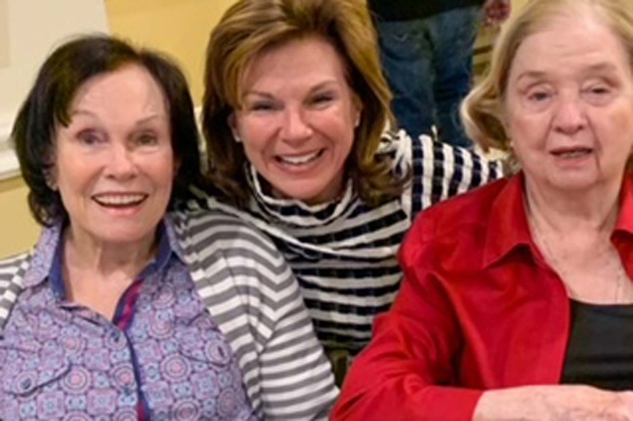 A group photo three women. The woman in the center has her arms around the shoulders of the two women on either side.