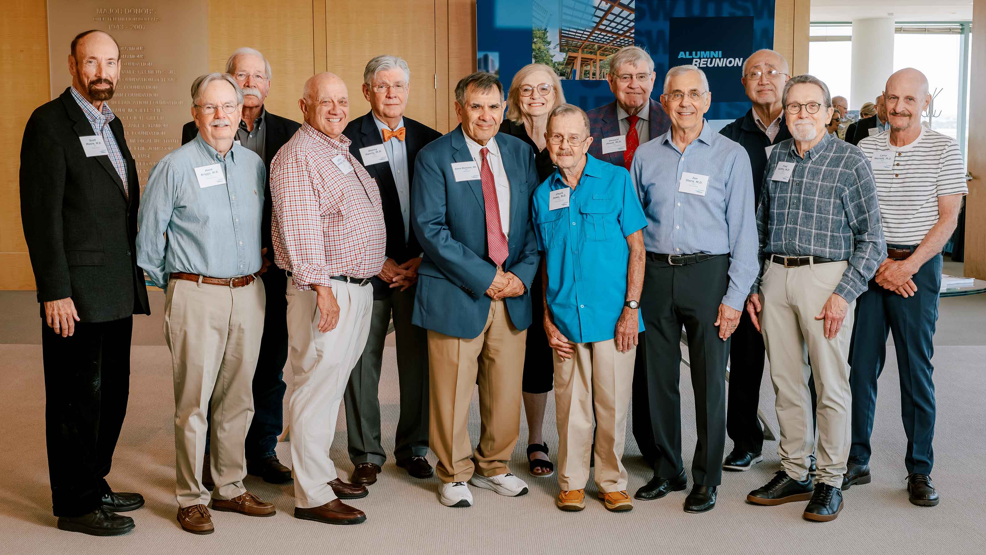 A group of 13 people standing in two rows in front of a banner that reads Alumni Reunion