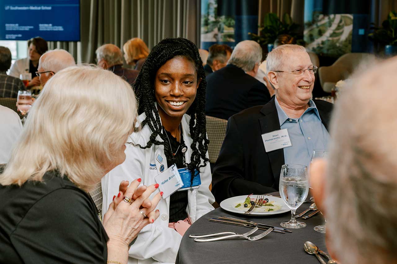 A female student wearing a lab coat and seated at a table listens to a woman speaking seated beside her.