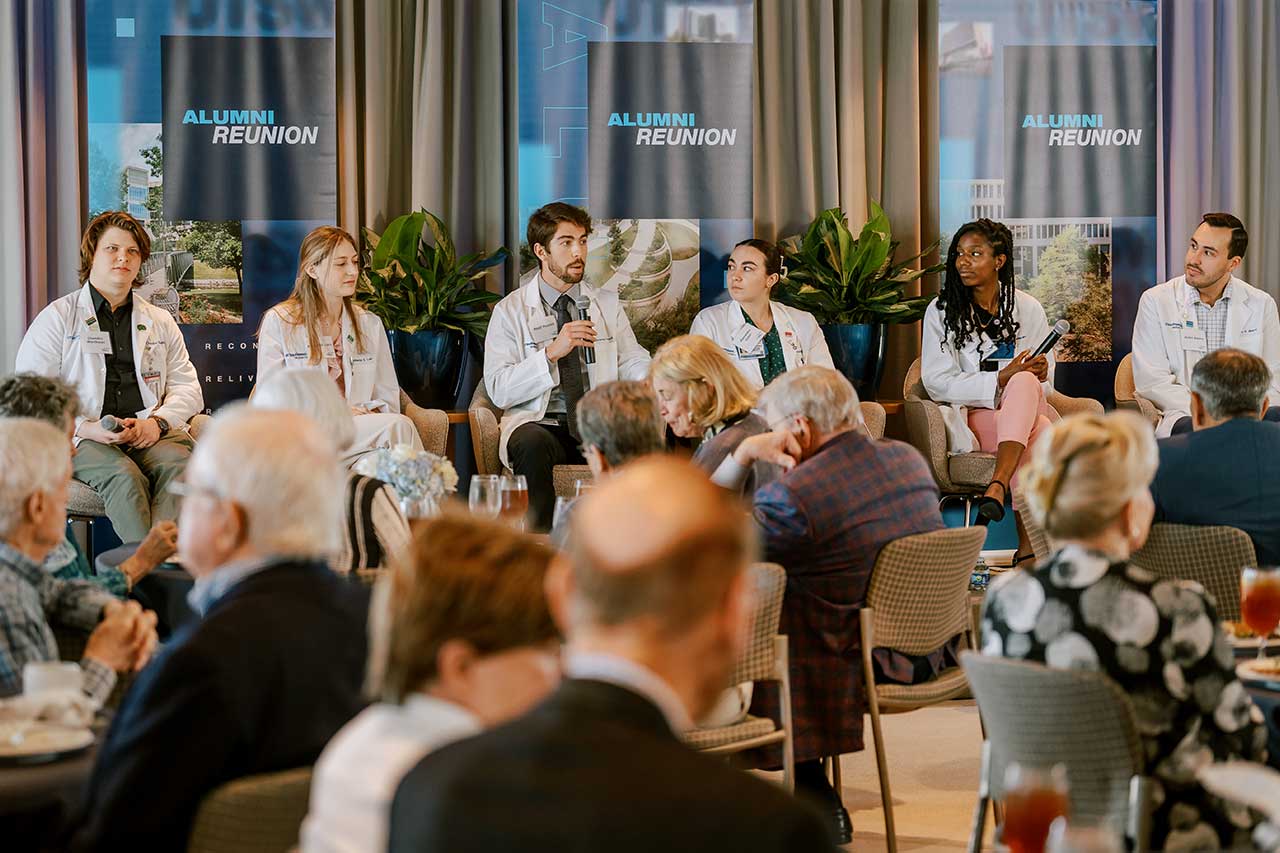 A male student seated in a chair on a stage with five other people and wearing a lab coat speaks to a room full of people seated at round tables.