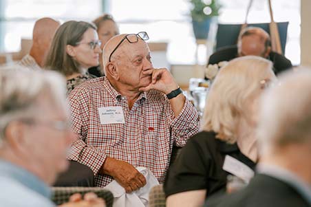 A man seated among a crowd of people rests his chin in his hand as he listens to a speaker.