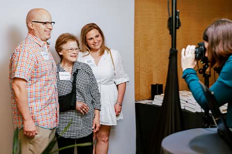 A man poses with two women against a photo backdrop as a photographer takes their photo.