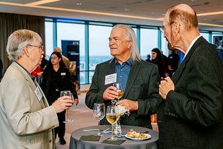 Three standing men chat at a cocktail table during a reception.