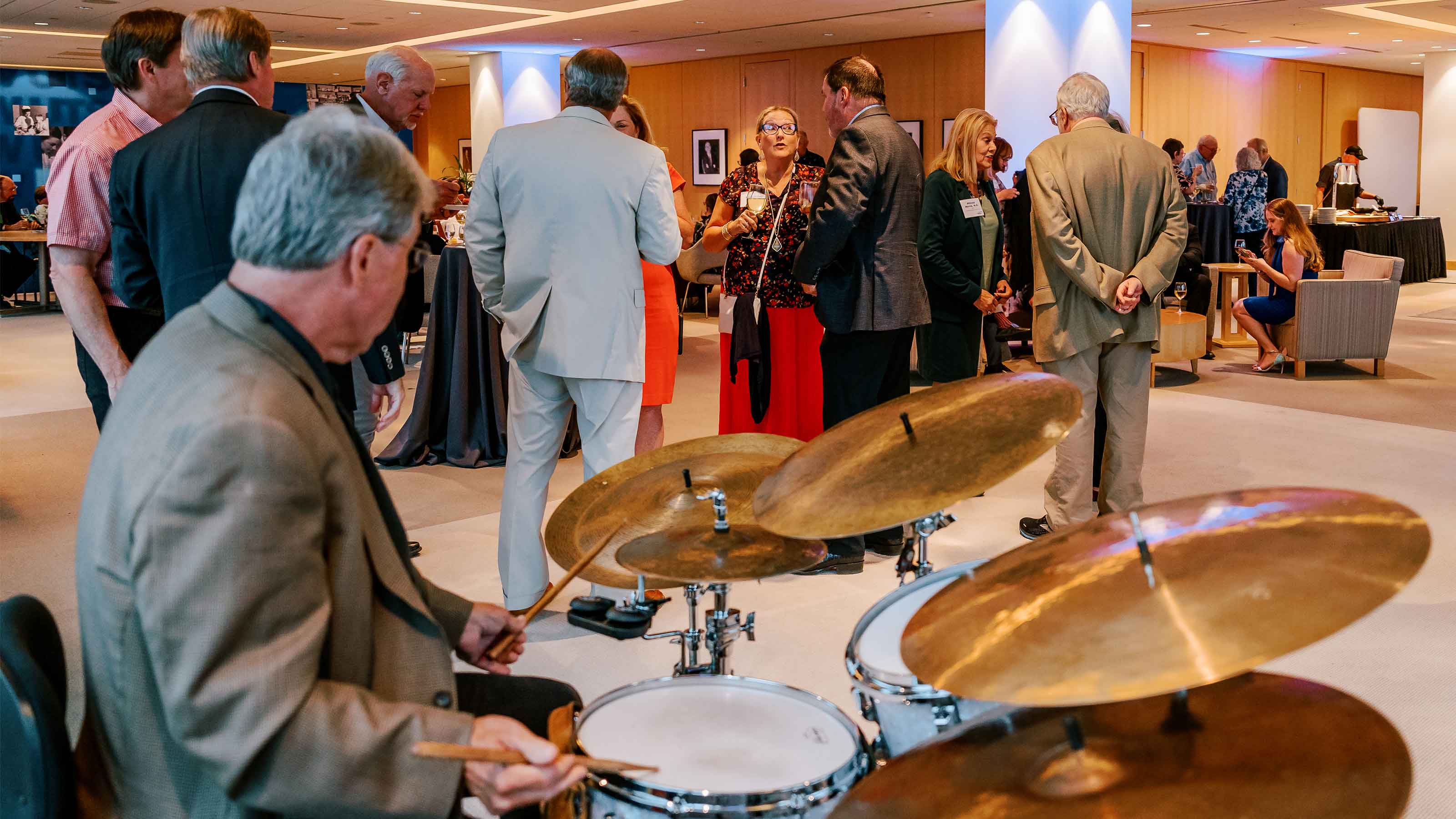 A drummer seated and playing a drumset looks out across a room of people chatting while standing and seated.