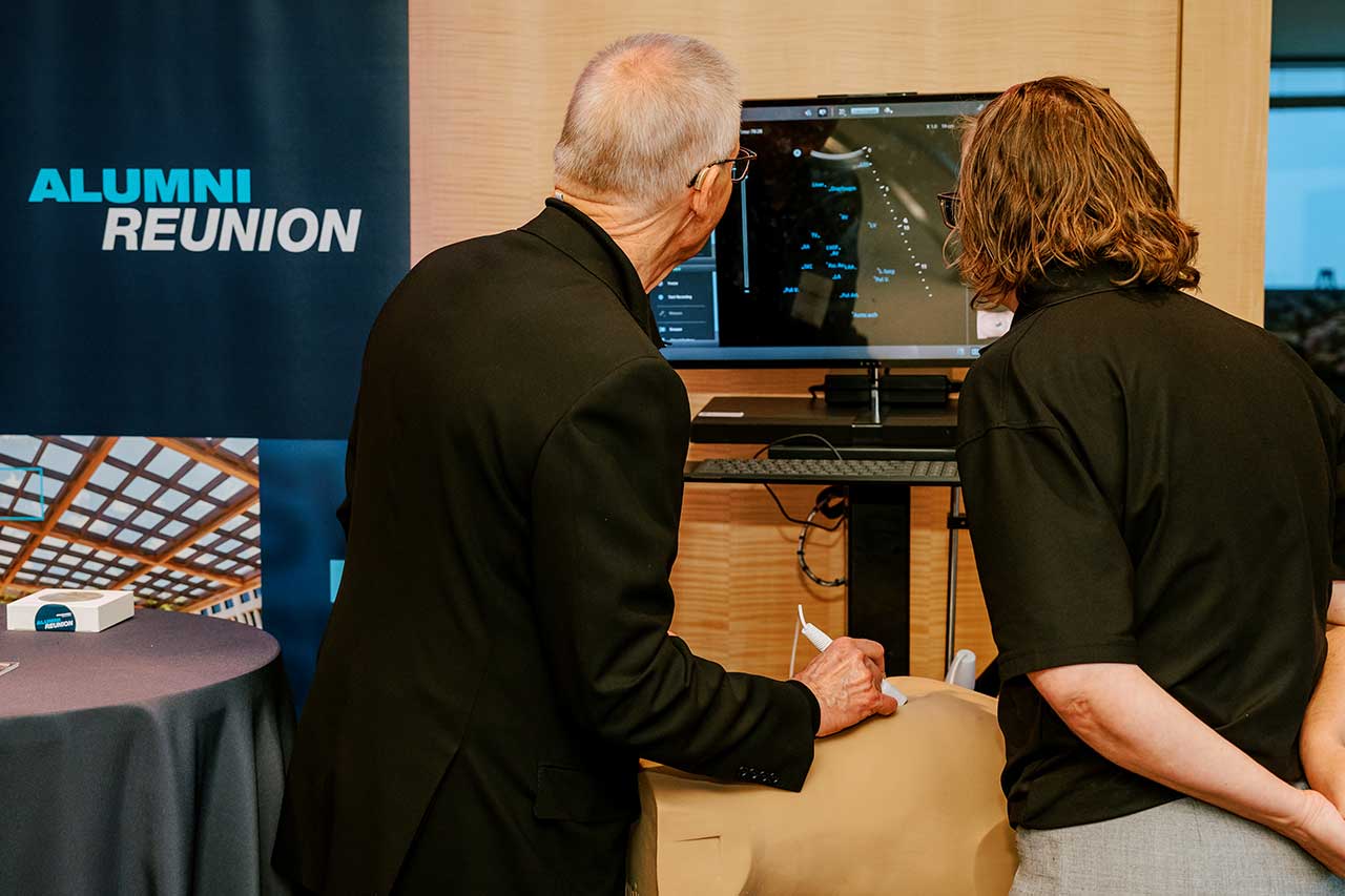 A woman stands beside a man who is holding an ultrasound tranducer against a simulation mannequin laying on a table in front of them. They both look at a computer monitor displaying medical imaging.