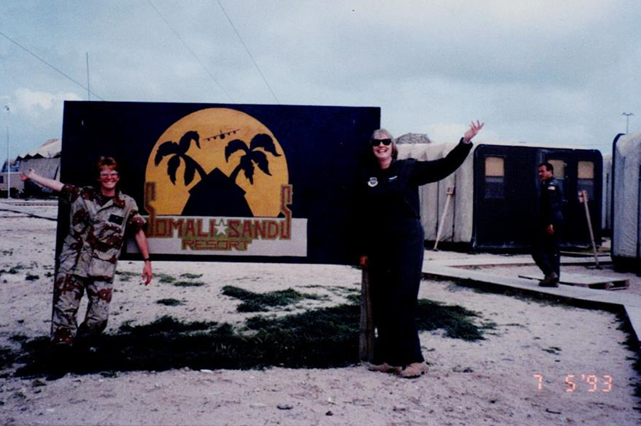 Gladys Young and another woman standing in a desert next to a sign that says 'Somalia Sands Resort'.