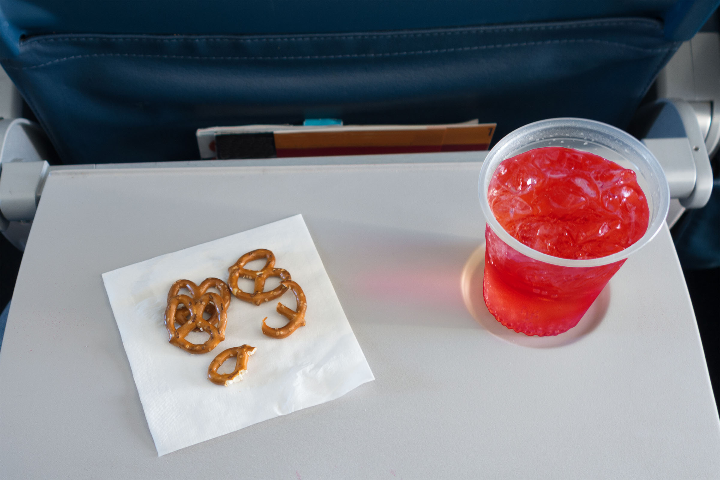 a first-person view of a few pretzels on a napkin and a red drink on an airplane eating tray.