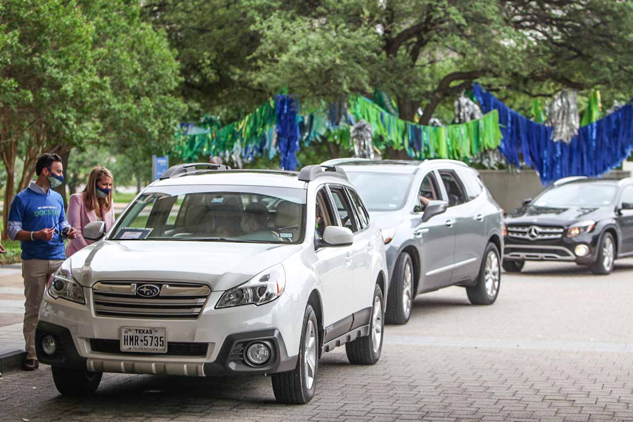 A line of cars drive onto the UT Southwestern campus past trees decorated with colorful streamers